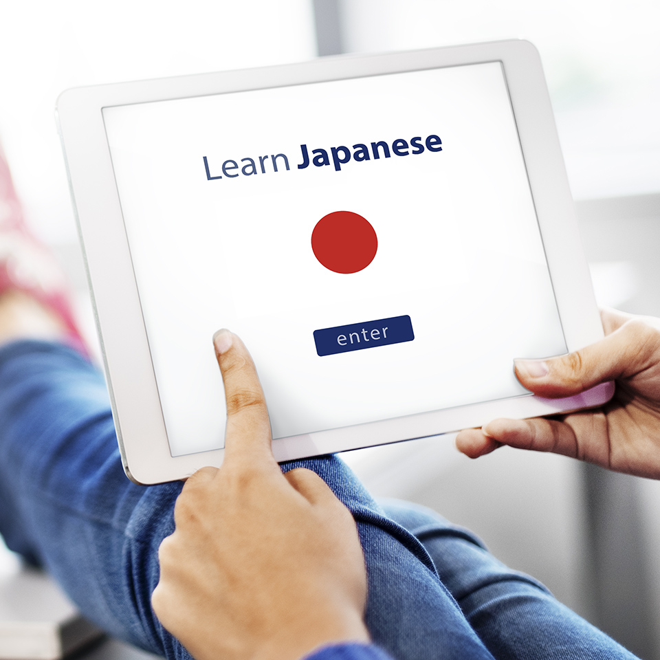 Hands holding up a tablet computer and touching the screen, which reads 'Learn Japanese - Enter' and displays the flag of Japan