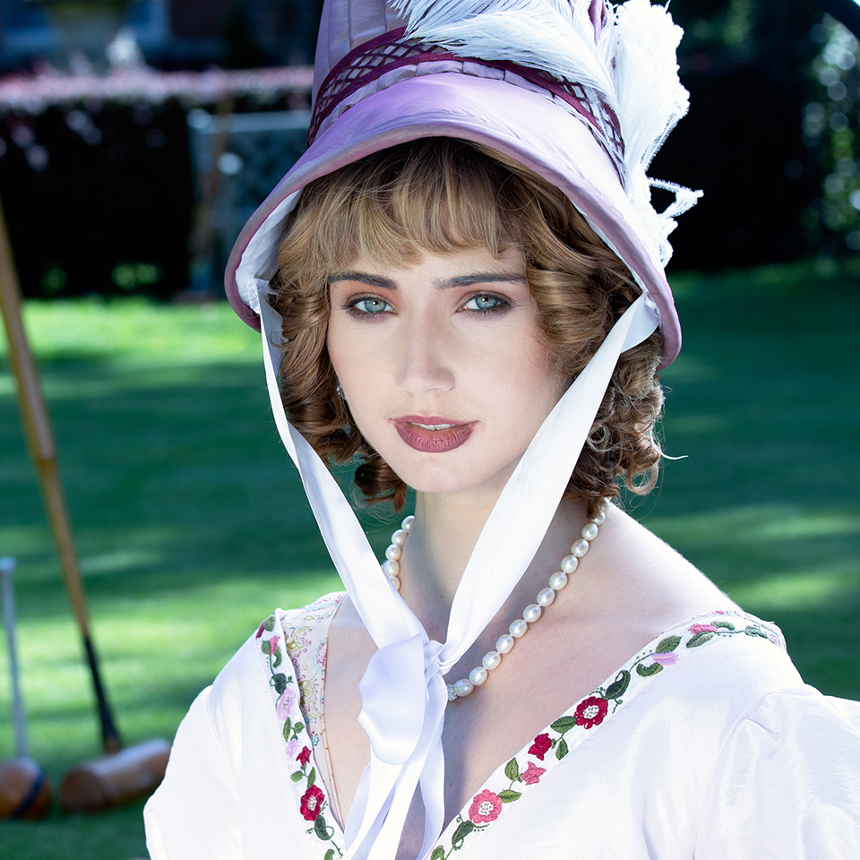 Woman in a vintage dress and bonnet sitting in front of a mansion with croquet sticks in the background