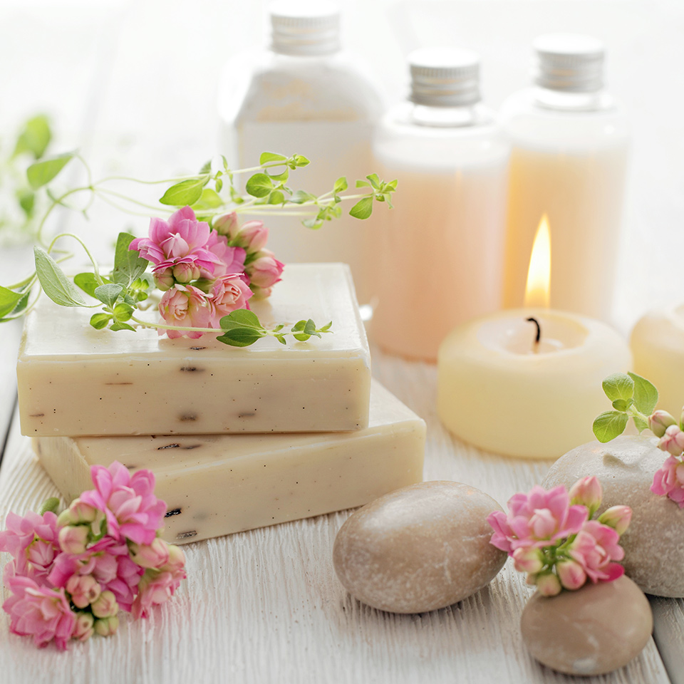 Aromatherapy products laid out on a wooden table. These include soap, candles, and lotions. Flowers and smoothed pebbles are also visible in the image.
